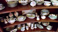 Full view of Royal Vienna china items on shelving including plates, teacups, pitchers, salt and pepper shakers, serving bowls and dishes all with floral and gold designs.