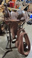 Front angle of brown tooled leather Western saddle mounted on folding work table with two harness collars hanging