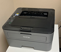 Front and top view of Brother HL-L2320D monochrome laser printer in gray and black casing on a white table.