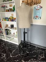 Full view of cloth-covered mannequin torso mounted on tall black wooden adjustable stand with tripod base, showing overall condition and surroundings.