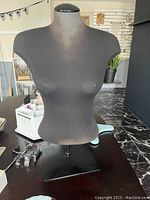 Front view of black fabric covered female mannequin bust with metal base on table, used for displaying clothes/jewelry