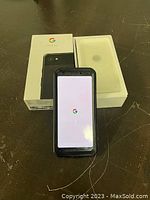 Google Pixel 3A phone powered on showing Google startup screen, next to original box and packaging.