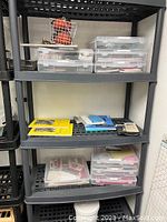Photo of grey resin freestanding shelving unit with 5 shelves displaying various clear plastic storage containers and office materials not included with lot.