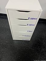 Front view of the white Ikea Alex cabinet showing five drawers with removable vinyl labels 'Paper', 'Glue', 'Painting', and 'Cleaning'.