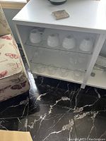 Close up of lower glass shelves inside white Ikea Besta unit, showing white ceramic cups and clear drinking glasses.