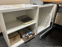 Frontal view of white Ikea Besta cabinet with shelf and storage inside containing paper and boxes