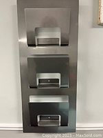 Front view of the stainless steel magazine rack mounted on wall showing three curved compartments for magazines.