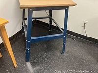 Corner view of metal tool stand with blue metal legs and light wood removable top on black floor with nearby wooden furniture and wall outlet