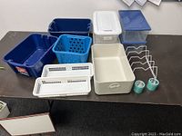 Various used plastic storage bins, baskets, and containers in multiple colors and sizes along with metal wire racks and small glass jars.