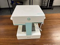 White and teal Mold Press machine on wooden table, front view with power button visible