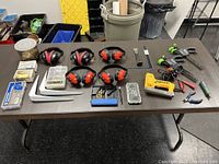 Wide view of table display showing ear protection earmuffs, cordless drill, clamps, drill bit sets, pliers, screws, and shelf brackets arranged neatly.