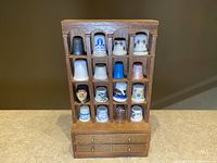 Photo showing 16 collectible thimbles in wooden case with three drawers, various ceramic and metal thimbles with decorative motifs