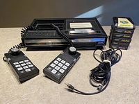 ColecoVision console with two controllers, power adapter cable, and six game cartridges stacked upright.