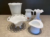 Full view of five milk glass items on countertop including vases, pitcher, basket, and top hat candy dish.