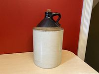 Front view of a large antique crock jug with light gray ceramic body and dark brown glossy top with handle, corn cob stopper visible at the opening.