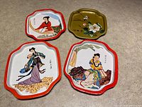 Four vintage metal snack or decorative plates displayed on a flat surface, showing the colorful Asian-inspired designs on three plates and floral design on the green plate.