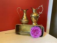 Front and side view of two brass decanter vases placed inside a rectangular brass planter with a purple flower in front.