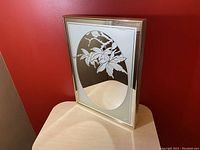 Front angled view showing the frosted floral artwork and oval mirror inside light wood frame, resting on a wooden surface with red wall behind.