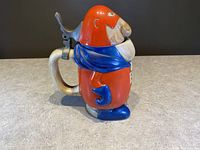 Side view of the vintage Bud Man stein showing the red, blue, and silver ceramic body with metal modal hinge lid and curved handle.