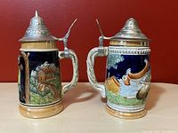 Two vintage West German beer steins showing side views. Left stein has pastoral cow and barn scene, right stein has male figures. Both have ornate pewter lids and cream-colored handles and base against deep blue background.