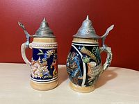 Two vintage West German beer steins with decorative scenes and pewter lids, shown from a front angle.