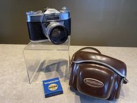 Voigtländer Bessamatic camera displayed on a clear stand with the lens attached, alongside a brown leather carrying case with Voigtländer logo and a Voigtländer branded filter box placed nearby.