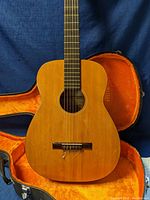 Front of vintage Harmony acoustic guitar showing natural wood body and bridge with nylon strings, in open orange-lined hard case.
