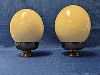 Front view of two ostrich egg sculptures on metal stands against a blue fabric background.