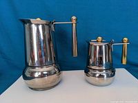 Front and side view of two stainless steel Italian espresso makers with gold-accented handles and knobs, placed on a white surface against a blue background.