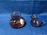 Front view of two purple translucent glass bird figurines with clear glass beaks and tails on a blue fabric background