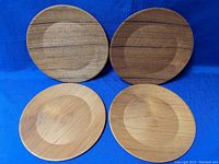 Four round wooden plates made of teak displayed on blue fabric background. Plates show natural wood grain and smooth finish.