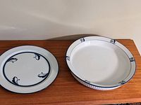 Two white ceramic pieces by Dansk on a wooden surface, showing a round plate and a pie plate with blue floral rim designs.