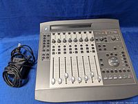 Full top view of the Digidesign Command 8 control surface showing 8 motorized faders, rotary knobs, buttons, and power supply cable next to it on a blue background.