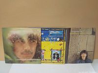 Three George Harrison vinyl LP records shown front covers: close-up portrait, colorful abstract yellow-themed art, George seated by stone wall.