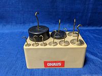 Front angled view of 9 OHAUS precision machined weights with hooks on a beige base.