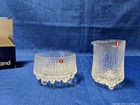 Front view of the glass cream and sugar set on blue fabric background, showing both pieces with the Iittala red dot logo.