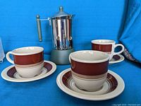 Photo showing three Norwegian-style cups and saucers with brown and beige bands, arranged around a stainless steel Italian stovetop coffee maker with a green handle and base.