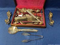 Photo of the open wooden box showing a large assortment of silverplated cutlery including knives, forks, spoons, and serving utensils on a red velvet lining.