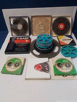 Photo showing mixed lot of reel to reel magnetic tape reels in original boxes and loose reels of various sizes and colors including blue plastic reels, black reels, and tape in cardboard sleeves