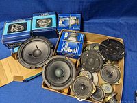 Lot showing multiple tweeters and midrange drivers packed in original boxes and loose units, displayed on cardboard and blue cloth background.