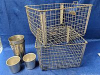 Two vintage metal wire baskets stacked with four metal cups beside them on a blue fabric background.