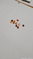 Image showing a collection of small, round cabochon orange carnelian gemstones scattered on white surface, varying shades of orange and red.