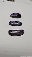 Three polished purple charoite stones displayed on a white surface, showing various shapes and unique natural patterns.