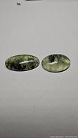Pair of oval cabochon cut natural mossy green rutilated prehnite stones on a white background, showing translucent greenish color with black inclusions.