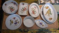 Top-down photo showing nine assorted Royal Worcester Evesham dishes on a woven mat, featuring bowls, platters, and bakeware with fruit decorations.
