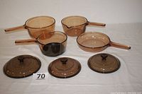 Four pieces of amber Vision Corning France glass cookware: 1.5L pot, 1L pot with spout, 1L pot with spout and black non-stick bottom, 7-inch saucepan. All with handles and lids shown.