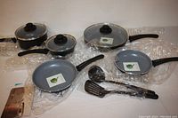 Full set of seven cookware pieces including three fry pans, two saucepans with lids, nylon spoon and turner, displayed wrapped in protective plastic.