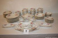 Full set of 84-piece porcelain china displayed on a table showing serving platters, tureen, gravy boat, plates, bowls, cups, and saucers with floral design and gold edges