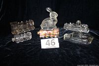 Six clear glass candy containers including railroad locomotives, a cabin cruiser boat, fire engine, and a rabbit arranged on black fabric with a lot number card.