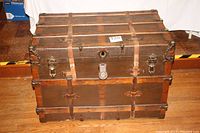 Closed large antique sea chest with metal reinforcements and leather straps, showing front and side metal latches, and broken leather handle.